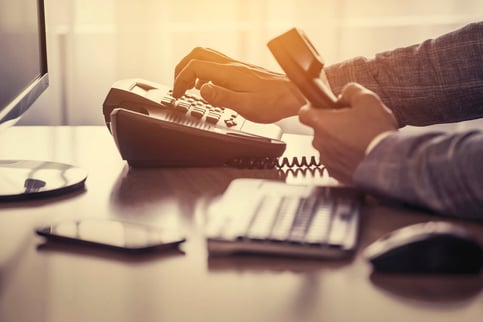 Do You Use an iPhone 3? Then Why Do You Use Old Business Phones? Employee holding a phone receiver in one hand and dialing out on their office desk phone with the other. Also visible is a keyboard, mouse, cell phone, and monitor.