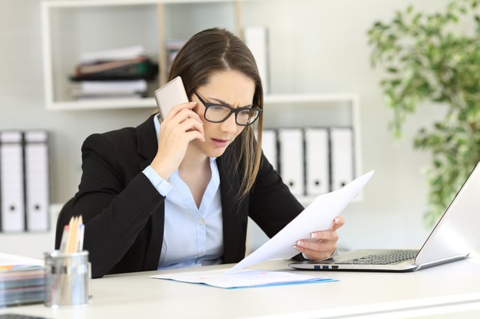 Business woman on a cell phone with a laptop open looking at a piece of paper.